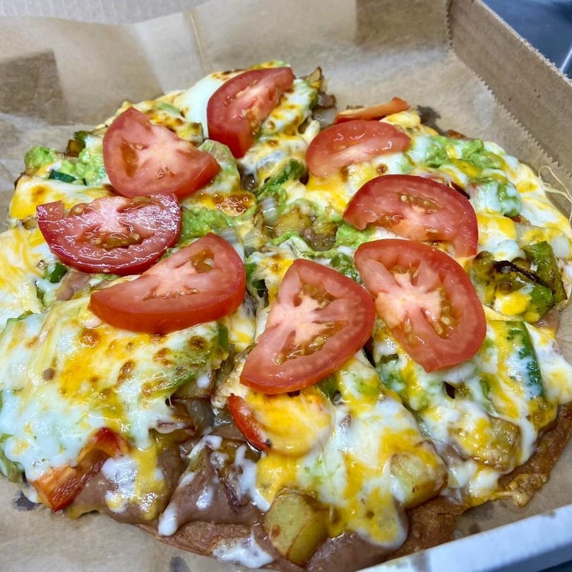 A round, cheesy pizza topped with sliced tomatoes, avocado, and other vegetables in a box.