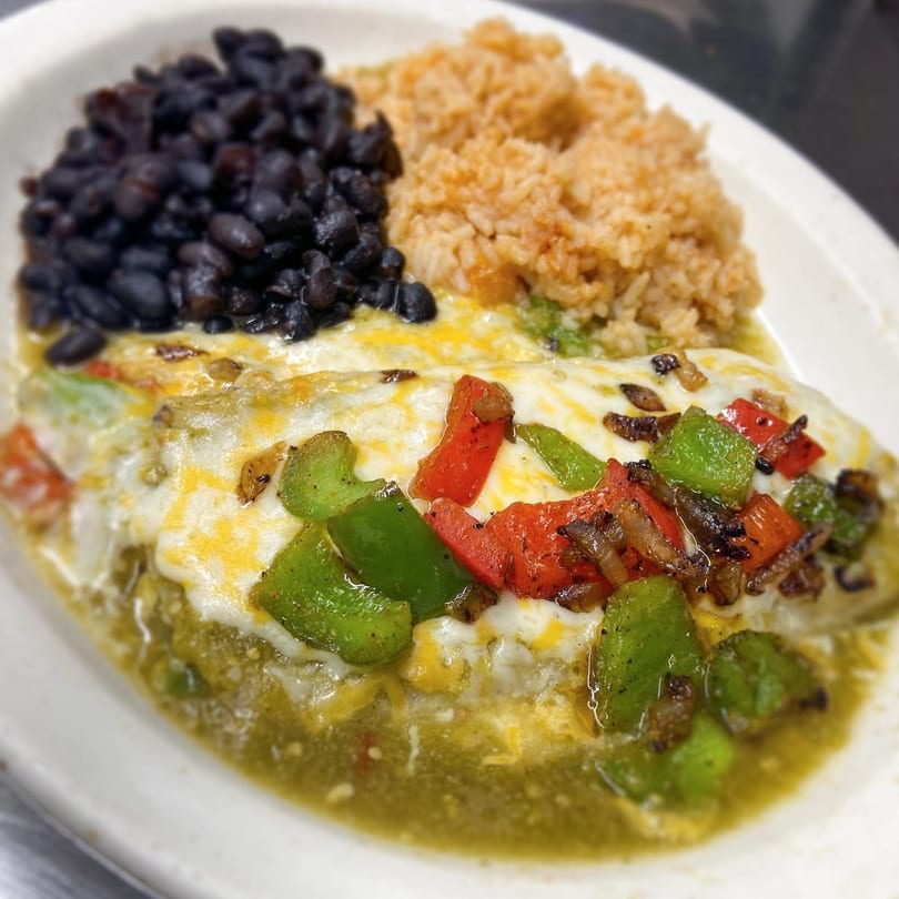 A plate of enchiladas topped with melted cheese, green and red peppers, served with black beans and rice.