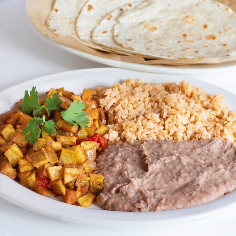 A plate of Mexican food with seasoned potatoes, rice, refried beans, and tortillas in the background.